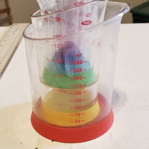 May include: A set of five clear plastic measuring cups with red markings. The cups are nested inside each other and have a red base. The cups are marked in milliliters (ml) and have a capacity of 250 ml.