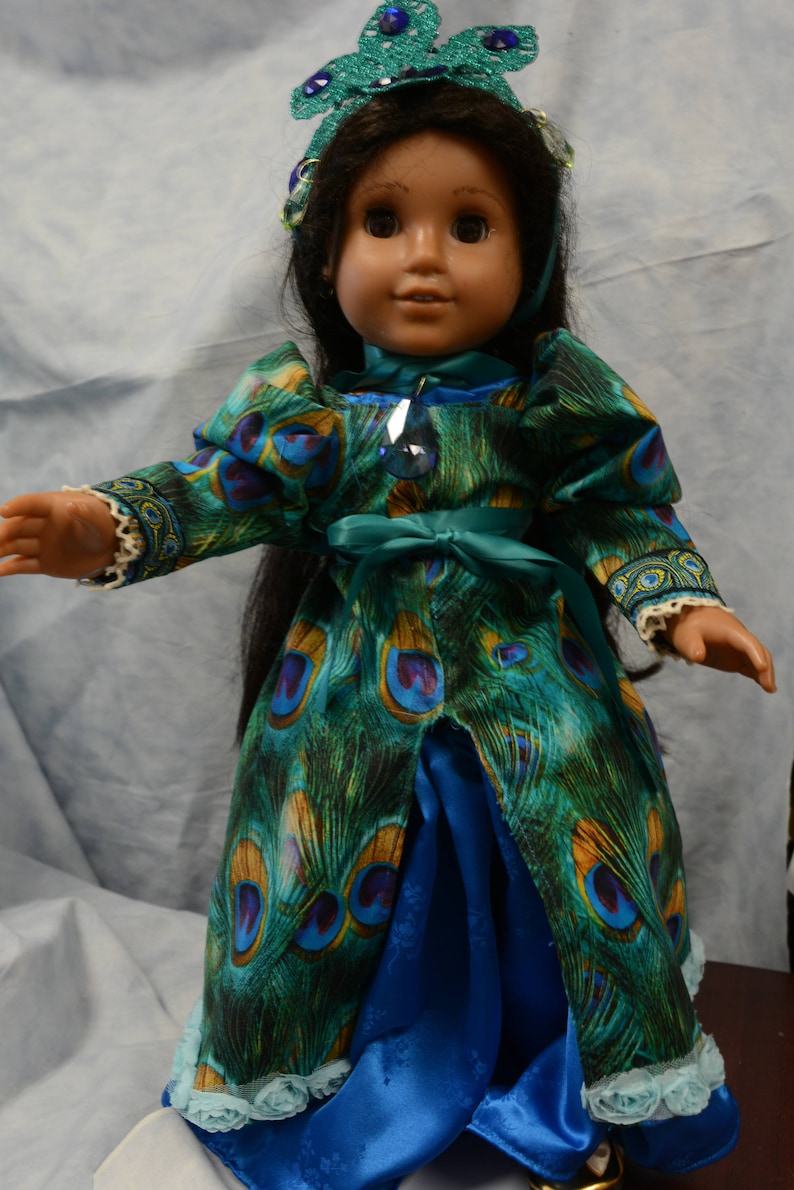 May include: A doll wearing a blue and green peacock-patterned dress with a blue satin skirt and a crown with blue jewels.