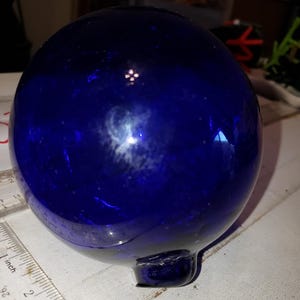 May include: A deep blue, spherical glass object with a small, rounded base. The glass has a glossy finish and reflects light, creating highlights. A ruler with inch measurements is in the foreground.