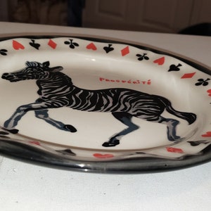 May include: A white ceramic plate with a black rim and a black and white zebra illustration. The plate is decorated with red hearts and black clubs around the rim. The text "Pansywild" is printed on the plate.