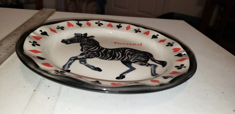 May include: A white ceramic plate with a black rim and a black and white zebra illustration. The plate is decorated with red hearts and black clubs around the rim. The text "Pansywild" is printed on the plate.