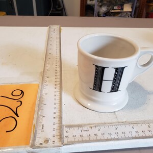May include: A white ceramic mug with a black letter "H" on the front. The mug has a handle and a wide base. A ruler is next to the mug, showing inch measurements.