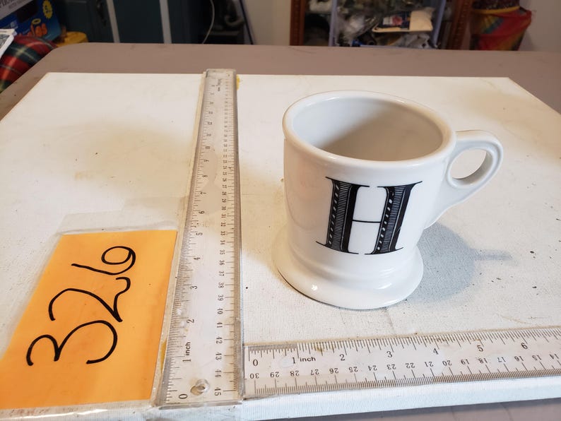 May include: A white ceramic mug with a black letter "H" on the front. The mug has a handle and a wide base. A ruler is next to the mug, showing inch measurements.
