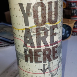 May include: A white travel mug with a world map design and the text "You are here" printed on it. The mug has a black lid and a black base.