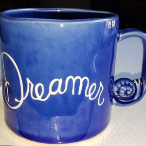 Pu&ograve; includere: Una tazza in ceramica blu con un manico bianco e la parola "Dreamer" scritta in bianco sul davanti.