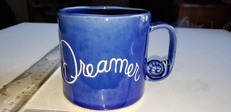Pu&ograve; includere: Una tazza in ceramica blu con un manico bianco e la parola "Dreamer" scritta in bianco sul davanti.
