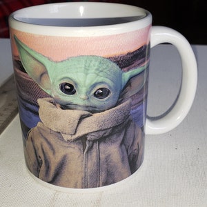 May include: White ceramic mug featuring a colourful image of a green character with large eyes, wearing a brown jacket and a brown scarf. The character is from the Star Wars franchise.