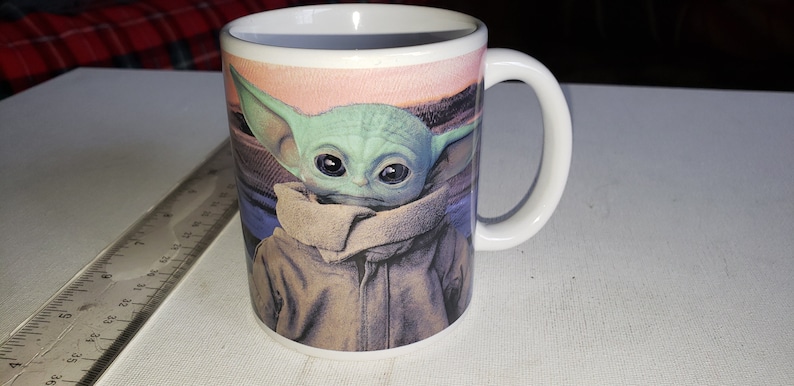 May include: White ceramic mug featuring a colourful image of a green character with large eyes, wearing a brown jacket and a brown scarf. The character is from the Star Wars franchise.