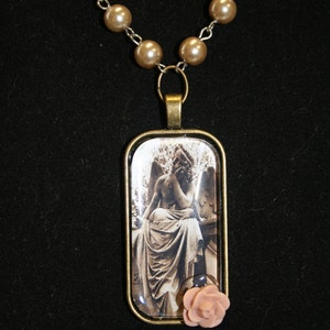 May include: A necklace with a brown rectangular pendant featuring a black and white image of a statue of an angel. The pendant has a pink flower charm attached to the bottom.