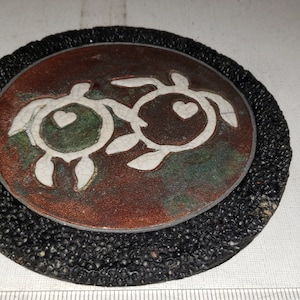 May include: A round coaster with a textured black border. The center features a copper-colored oval with two white turtle silhouettes, each with a heart. The turtles are outlined in white and have green accents.