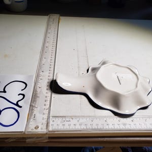 May include: A white ceramic spoon rest with a scalloped edge and a dark blue trim. The spoon rest is in the shape of a seashell. A ruler is next to the spoon rest. The number 825 is written on a piece of paper.