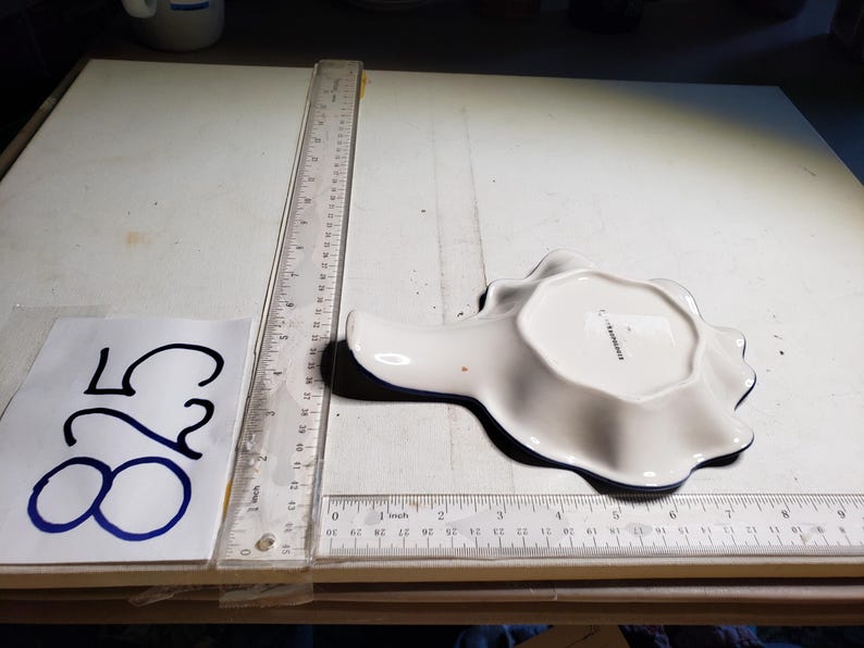 May include: A white ceramic spoon rest with a scalloped edge and a dark blue trim. The spoon rest is in the shape of a seashell. A ruler is next to the spoon rest. The number 825 is written on a piece of paper.