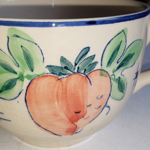 Puede incluir: Una taza de cerámica de color crema con un borde azul y un diseño pintado a mano de un melocotón con hojas verdes.