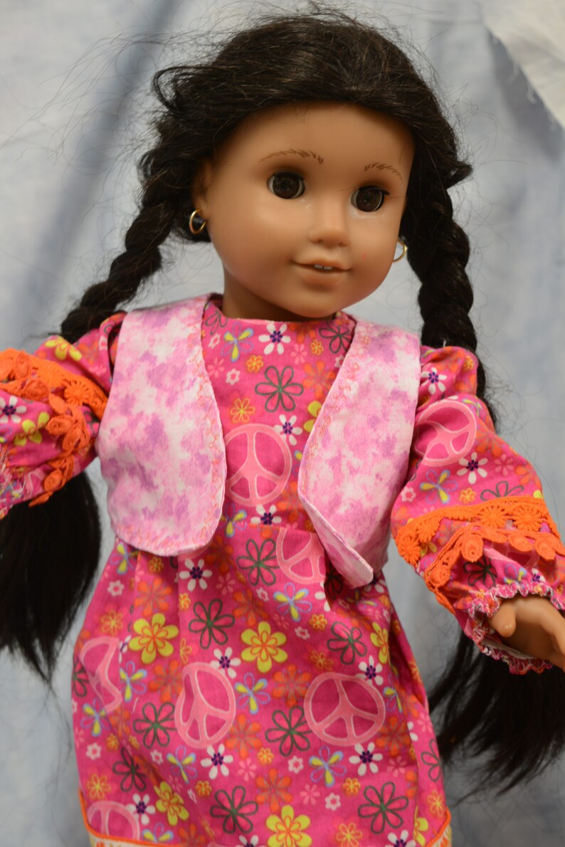 May include: A doll wearing a pink and white vest over a pink dress with a peace sign and flower pattern. The doll has long black hair in braids and brown eyes.