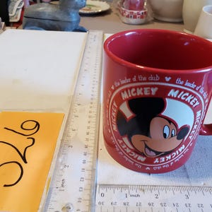 Puede incluir: Una taza de cer&aacute;mica roja con Mickey Mouse. La taza tiene un asa grande y un dise&ntilde;o circular con la cara de Mickey y las palabras "Mickey" y "the leader of the club". La taza est&aacute; junto a una regla y una etiqueta amarilla con el n&uacute;mero 326.