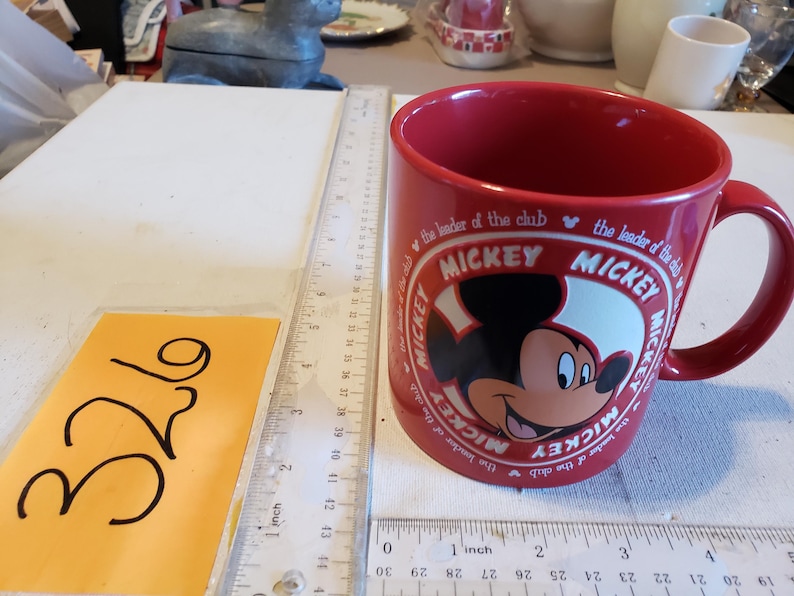 Puede incluir: Una taza de cer&aacute;mica roja con Mickey Mouse. La taza tiene un asa grande y un dise&ntilde;o circular con la cara de Mickey y las palabras "Mickey" y "the leader of the club". La taza est&aacute; junto a una regla y una etiqueta amarilla con el n&uacute;mero 326.