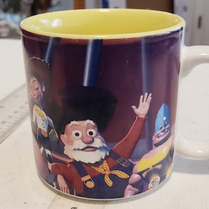 May include: A white ceramic mug with a yellow rim and a colorful image of characters from the movie Toy Story. The characters are Woody, Buzz Lightyear, and Jessie. The mug is a great gift for fans of the movie.