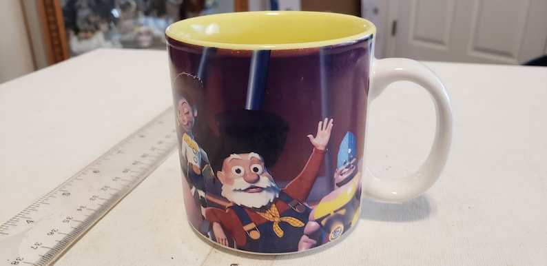 May include: A white ceramic mug with a yellow rim and a colorful image of characters from the movie Toy Story. The characters are Woody, Buzz Lightyear, and Jessie. The mug is a great gift for fans of the movie.