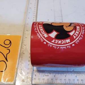 Puede incluir: Una taza de cer&aacute;mica roja con Mickey Mouse. La taza tiene un c&iacute;rculo blanco con la cara de Mickey y las palabras "MICKEY" y "the leader of the club" en un patr&oacute;n circular. El n&uacute;mero "326" est&aacute; en una nota adhesiva amarilla.