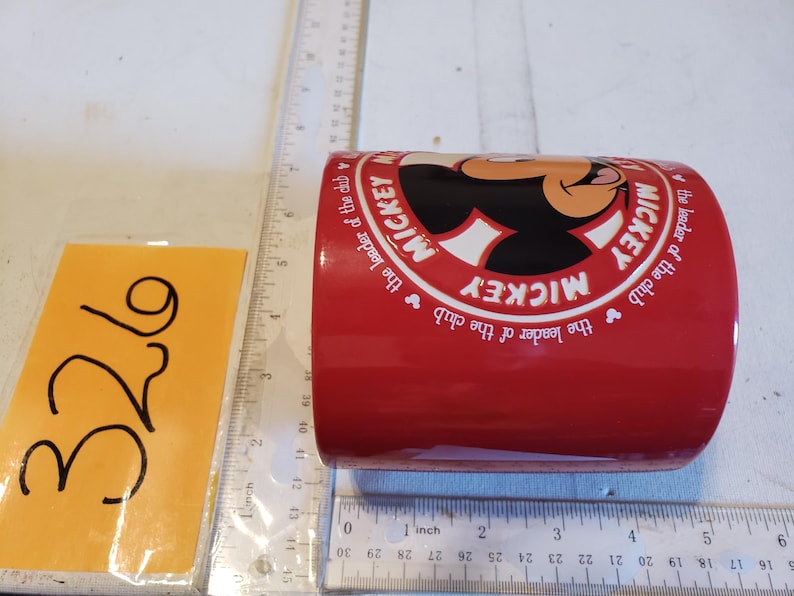 Puede incluir: Una taza de cer&aacute;mica roja con Mickey Mouse. La taza tiene un c&iacute;rculo blanco con la cara de Mickey y las palabras "MICKEY" y "the leader of the club" en un patr&oacute;n circular. El n&uacute;mero "326" est&aacute; en una nota adhesiva amarilla.