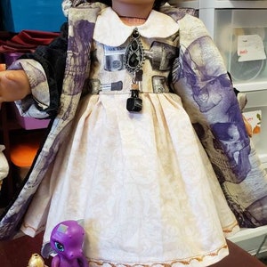 May include: A doll wearing a cream-colored dress with a floral pattern and a black and purple skull-print coat. The doll has dark hair and is wearing earrings. Accessories include a purple toy dragon and glass jars.