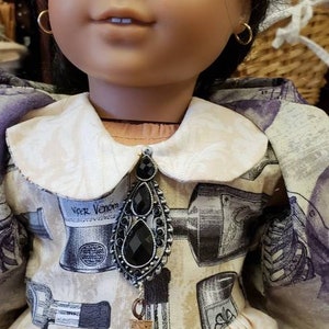 May include: A doll wearing a cream-colored dress with a white collar and a black pendant. The dress has a print of cosmetic items. The pendant is connected to a small black bottle with the text "EXTERNAL USE" on it.