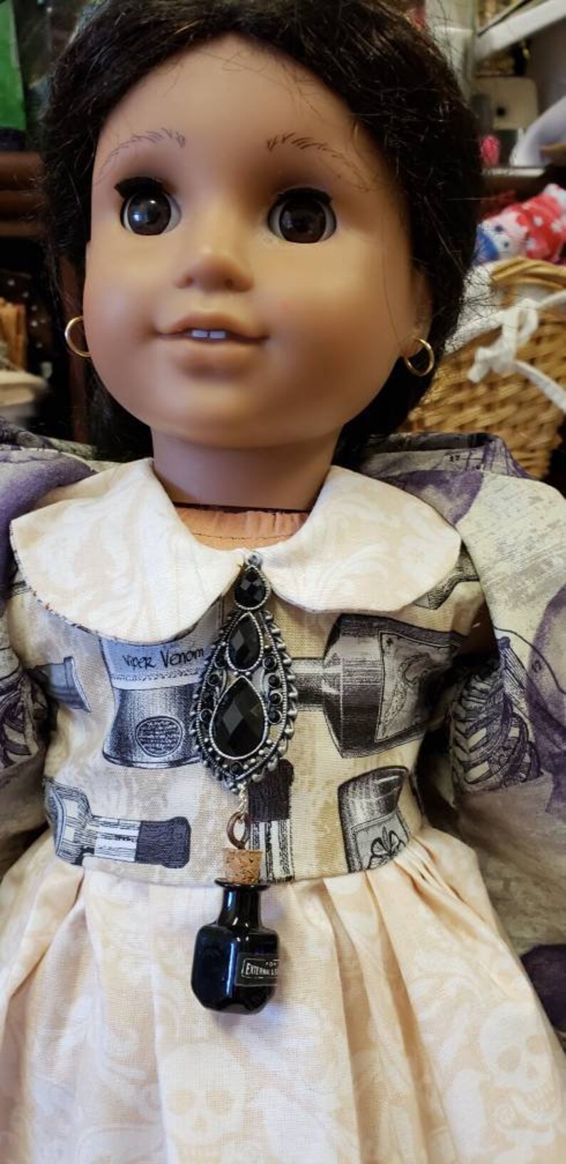 May include: A doll wearing a cream-colored dress with a white collar and a black pendant. The dress has a print of cosmetic items. The pendant is connected to a small black bottle with the text "EXTERNAL USE" on it.