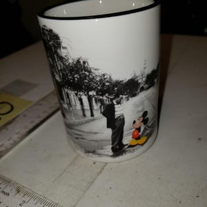 以下が含まれることがあります： 黒い縁取りのある白いセラミックマグカップ。マグカップには、ミッキーマウスと人が通りに立っている白黒写真が描かれています。ミッキーマウスは赤いショートパンツと黄色の靴を履いています。
