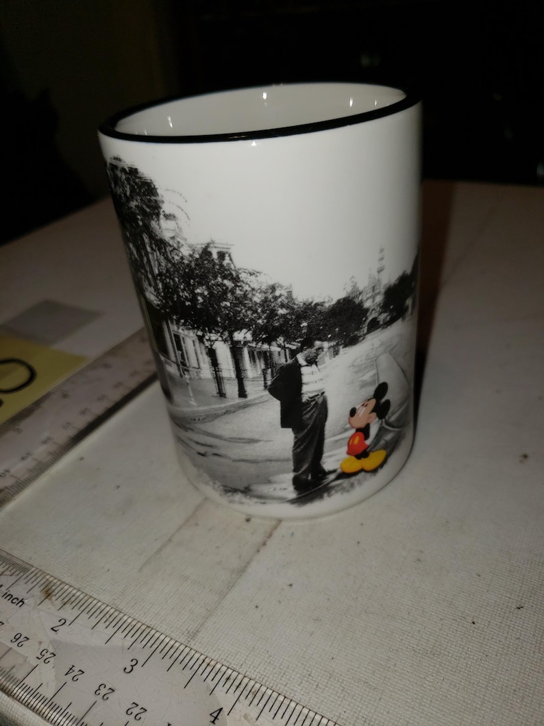 以下が含まれることがあります： 黒い縁取りのある白いセラミックマグカップ。マグカップには、ミッキーマウスと人が通りに立っている白黒写真が描かれています。ミッキーマウスは赤いショートパンツと黄色の靴を履いています。