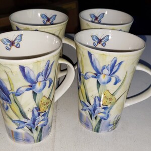 May include: Set of four ceramic mugs with a floral design featuring blue irises and butterflies. The mugs have a cream background with green leaves and yellow accents.