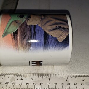 May include: White ceramic mug featuring a colourful image of a green alien character from the Star Wars franchise. The alien is wearing a brown hooded cloak and is standing on a desert planet.