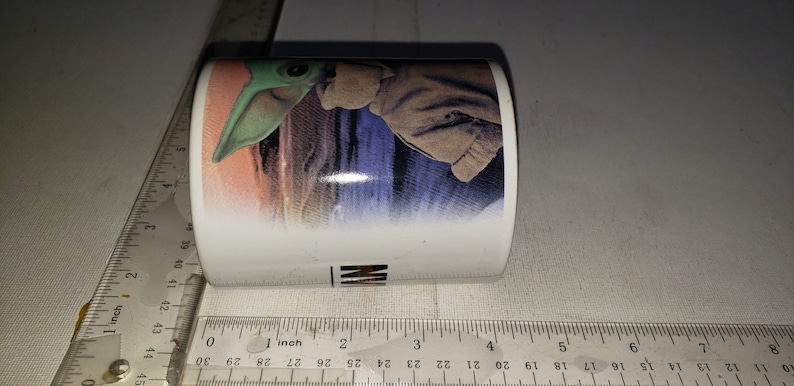 May include: White ceramic mug featuring a colourful image of a green alien character from the Star Wars franchise. The alien is wearing a brown hooded cloak and is standing on a desert planet.