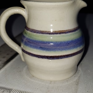 May include: A small, white ceramic pitcher with a blue, green, and brown striped design. The pitcher has a handle and a spout.