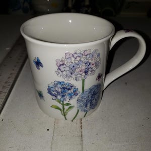 May include: White ceramic mug featuring a floral design of blue and purple hydrangeas and small blue butterflies. The mug has a curved handle and a smooth, glossy finish. The mug is approximately 4 inches tall.