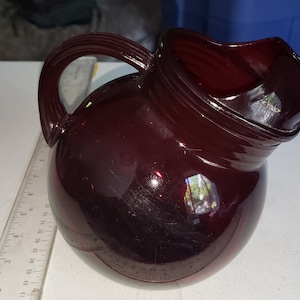 May include: A dark red glass pitcher with a rounded body and a wide spout. The pitcher has a smooth, glossy finish.