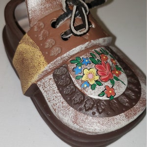 May include: A brown ceramic shoe with a white and brown painted design. The shoe has a yellow strap and a black leather lace. The shoe is decorated with a floral pattern in shades of blue, yellow, red, and green.