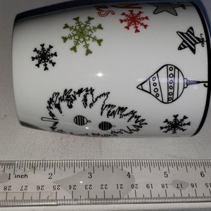 May include: White ceramic mug with a black and white Christmas design. The design features snowflakes, stars, and ornaments. The mug is approximately 3.5 inches tall.