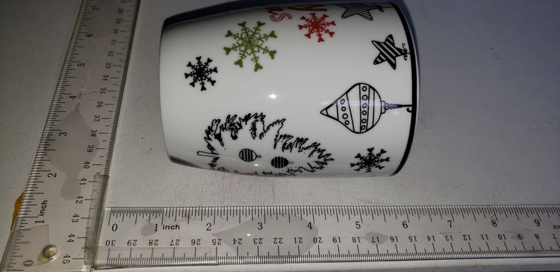 May include: White ceramic mug with a black and white Christmas design. The design features snowflakes, stars, and ornaments. The mug is approximately 3.5 inches tall.