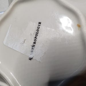 May include: White ceramic bowl with a scalloped edge and a blue trim. The bowl has a label that reads "by ANTHROPOLOGIE". The bowl is likely used for serving or display.