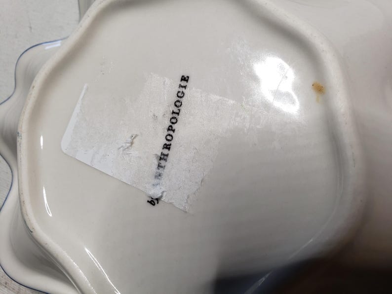 May include: White ceramic bowl with a scalloped edge and a blue trim. The bowl has a label that reads "by ANTHROPOLOGIE". The bowl is likely used for serving or display.