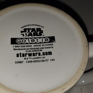 May include: White ceramic mug with a black Star Wars logo and Galerie branding. The mug has a black handle and the bottom of the mug has the text "starwars.com" and a phone number.