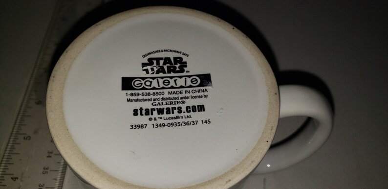 May include: White ceramic mug with a black Star Wars logo and Galerie branding. The mug has a black handle and the bottom of the mug has the text "starwars.com" and a phone number.