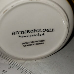 Op de afbeelding: Witte keramische mok met gele en zwarte strepen. De onderkant van de mok heeft de tekst "ANTHROPOLOGIE hand painted" en "DISHWASHER & MICROWAVE SAFE MADE IN CHINA/FABRIQU&Eacute; EN CHINE".