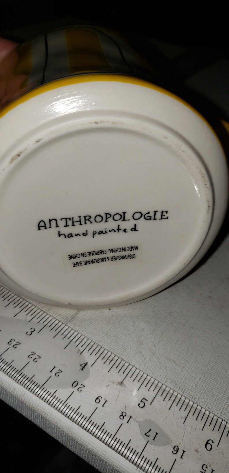 Op de afbeelding: Witte keramische mok met gele en zwarte strepen. De onderkant van de mok heeft de tekst "ANTHROPOLOGIE hand painted" en "DISHWASHER & MICROWAVE SAFE MADE IN CHINA/FABRIQU&Eacute; EN CHINE".