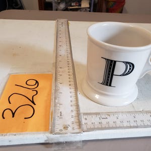以下が含まれることがあります： 黒い文字「P」のデザインが施された白いセラミックマグ。マグにはハンドルと広いベースが付いています。マグの隣には定規があり、「326」の数字が入った黄色のタグも表示されています。