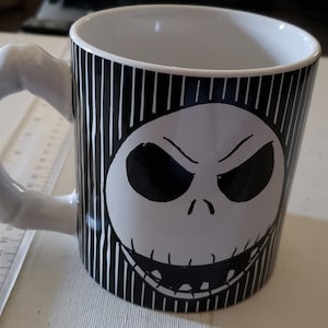 May include: A white ceramic mug with a black and white striped design. The mug features a black and white illustration of Jack Skellington's face from the movie The Nightmare Before Christmas.