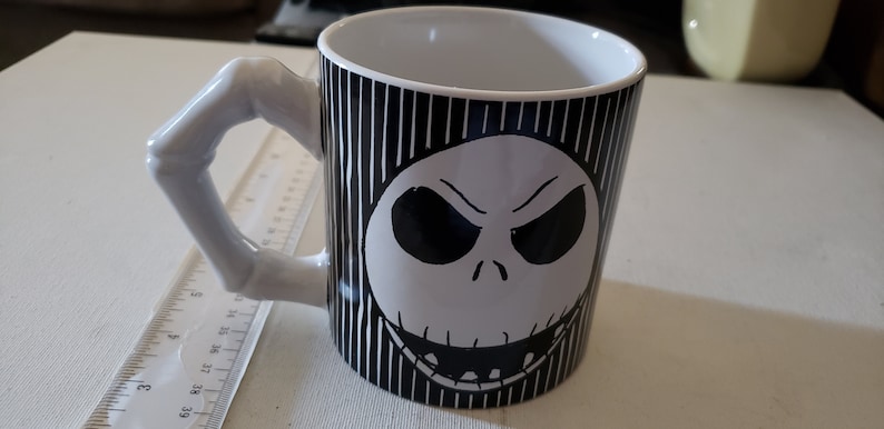 May include: A white ceramic mug with a black and white striped design. The mug features a black and white illustration of Jack Skellington's face from the movie The Nightmare Before Christmas.