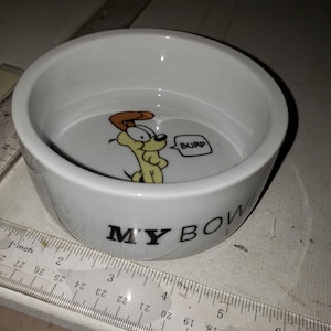 May include: White ceramic pet bowl with the words "MY BOWL" in black lettering. The bowl features a cartoon dog character with a speech bubble that says "BURP". A ruler is next to the bowl.