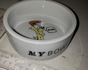 Puede incluir: Cuenco blanco de cerámica para mascotas con las palabras "MY BOWL" en letras negras. El cuenco presenta un personaje de perro de dibujos animados con un bocadillo que dice "BURP". Una regla está al lado del cuenco.