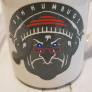 May include: White ceramic mug with a graphic of a grumpy face wearing glasses, with the words "BAH HUMBUG!" in a circular design. The face is gray with blue eyes and red-framed glasses. The mug has a handle on the right side.